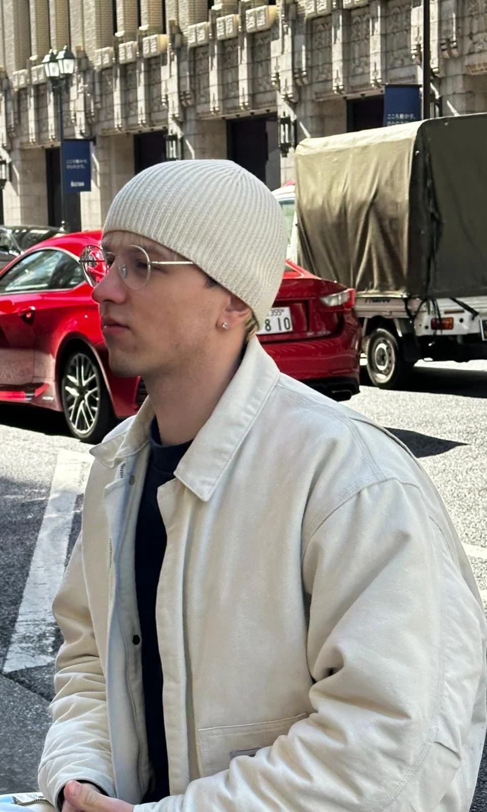 Junger Mann mit Brille, weißer Beanie und heller Jacke blickt auf einer Stadtstraße zur Seite.