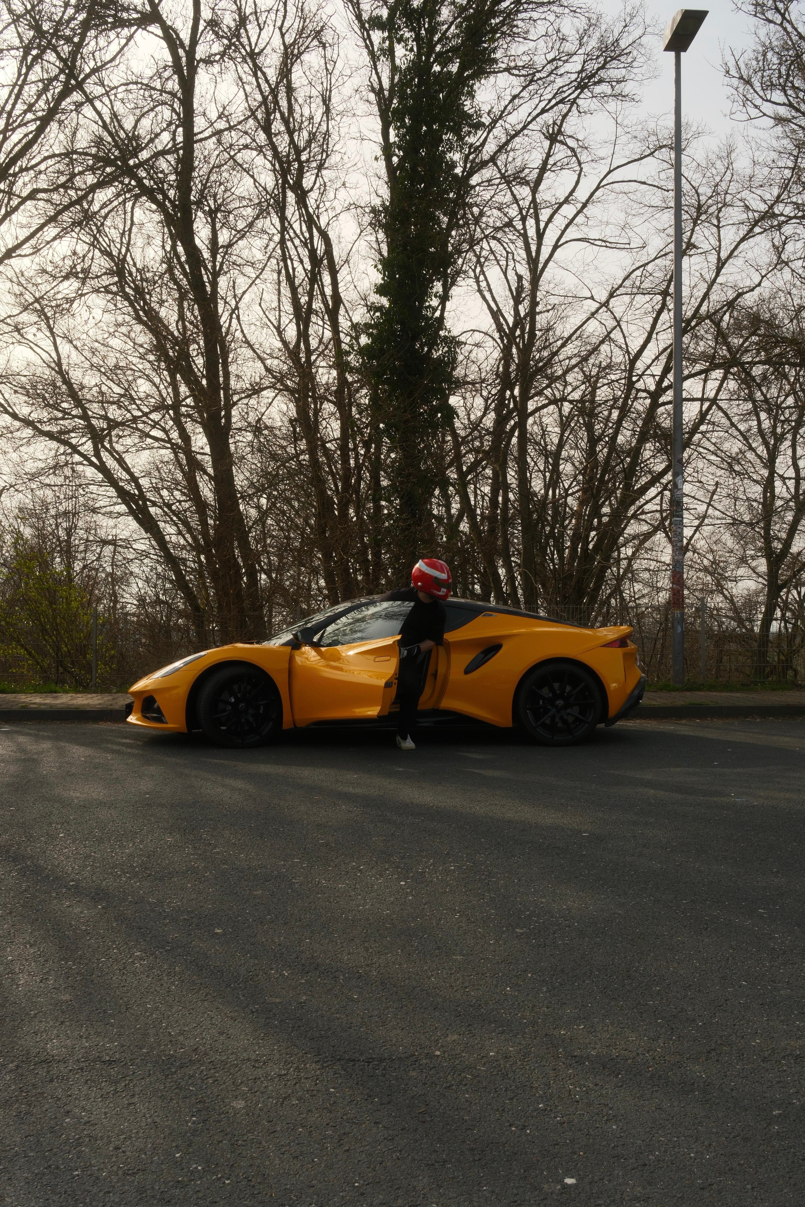 Gelber Sportwagen vor kahlen Bäumen, eine Person mit rotem Helm steigt aus der offenen Tür.