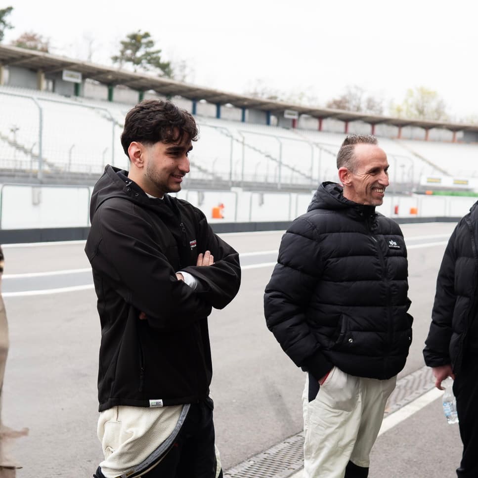 Zwei lächelnde Männer in schwarzen Jacken stehen auf einer Rennstrecke vor einer Tribüne.