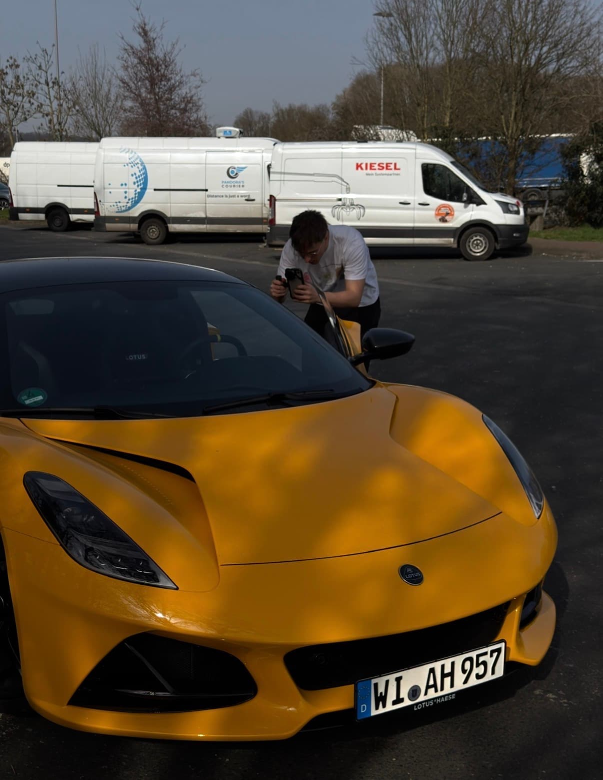Ein Mann fotografiert einen gelben Lotus Sportwagen auf einem Parkplatz vor zwei weißen Lieferwagen.